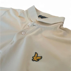 Beige pikétröja från Lyle & Scott - Snygg beige pikétröja från Lyle & Scott med klassisk krage och knappslå. Tröjan har ett broderat gult emblem på bröstet. Perfekt för en stilren och avslappnad look.