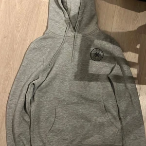 Grå hoodie med tryck från Tyskland - Säljer en grå hoodie med tryck från Tyskland. Den har en stor ficka framtill och ett tryck med texten 'Germany' och 'Deutscher Fussball Bund' på ryggen. 