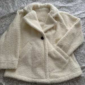 Vit teddyjacka/kappa - Mysig vit teddyjacka med en knapp framtill. Perfekt för kyliga dagar med sin fluffiga och varma design. Jackan har en klassisk krage och två fickor på sidorna. Den är fluffig och varm innuti också. Oanvänd