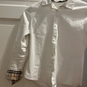 Vit skjorta från Burberry - Snygg vit skjorta från Burberry med klassiskt rutigt mönster på insidan av kragen och ärmsluten. Skjortan har långa ärmar och knappar framtill. Perfekt för en stilren look.