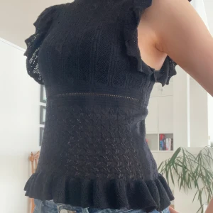 Svart topp med volangdetaljer - Snygg svart topp med volangdetaljer på ärmarna och nederkanten. Toppen har en ribbad struktur och är ärmlös, vilket ger en elegant look. Perfekt för att piffa upp din garderob! från zara