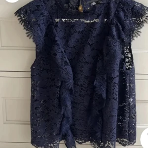 Mörkblå spetsblus från Zara - Elegant mörkblå spetsblus från Zara med vackra volangdetaljer. Blusen är ärmlös och har en fin struktur som ger en lyxig känsla. Perfekt för att lyfta vilken outfit som helst.