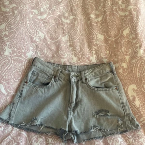 Grå jeansshorts med slitningar - Säljer ett par grå jeansshorts med coola slitningar och fransiga kanter. Shortsen har klassisk femficksdesign och knappgylf.