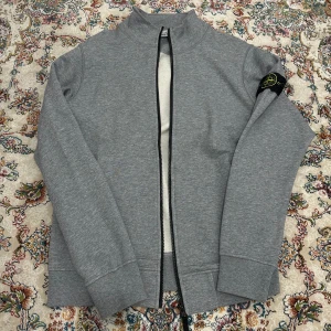 zip-tröja från Stone Island - Snygg grå zip-tröja från Stone Island med deras ikoniska märke på ärmen. Tröjan har en dragkedja framtill och är tillverkad i ett mjukt material. Perfekt för en stilren och avslappnad look.