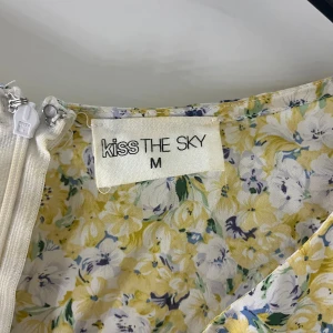 Blommig klänning från Kiss The Sky - Charmig blommig klänning från Kiss The Sky i storlek M. Klänningen har ett gult och vitt blommönster med långa ärmar och en knytning framtill. Den har en öppen rygg och en dragkedja för enkel påklädning.