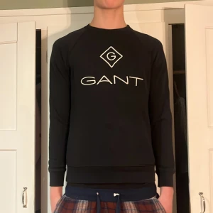 Svart sweatshirt från GANT - Snygg svart sweatshirt från GANT med stort tryck på framsidan. Tröjan har långa ärmar och en klassisk rund halsringning. Perfekt för en avslappnad stil.