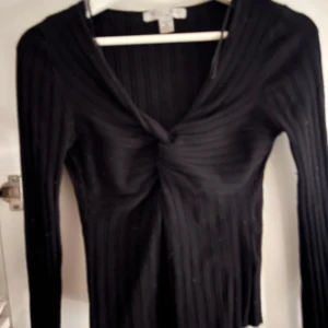 Svart ribbad tröja med knutdetalj - Snygg svart ribbad tröja med en unik knutdetalj framtill. Tröjan har långa ärmar och en v-ringad design som ger en stilren look. Perfekt för att klä upp eller ner beroende på tillfälle.