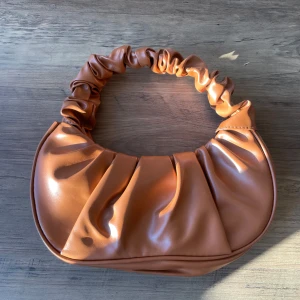 Brun axelväska i syntet - Snygg brun axelväska i syntetmaterial med rynkad axelrem. 