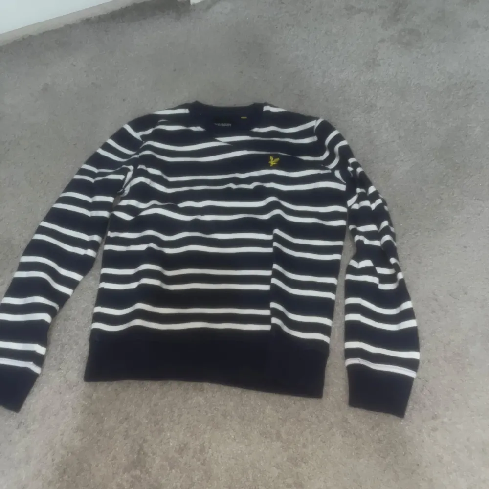 Snygg randig tröja från Lyle & Scott i blått och vit med ett gult broderat emblem på bröstet. Tröjan har långa ärmar och en klassisk passform. Perfekt för en stilren look.. Neuletakit & Villapaidat.