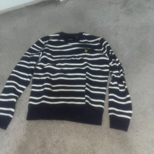 Randig tröja från Lyle & Scott - Snygg randig tröja från Lyle & Scott i blått och vit med ett gult broderat emblem på bröstet. Tröjan har långa ärmar och en klassisk passform. Perfekt för en stilren look.