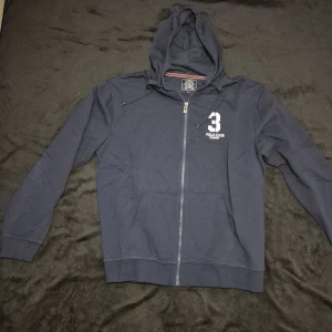 Mörkblå hoodie från Polo Club - Säljer en mörkblå hoodie från Polo Club med dragkedja och nummerdetalj på bröstet. Tröjan har en klassisk design med huva och långa ärmar. Perfekt för en avslappnad stil.