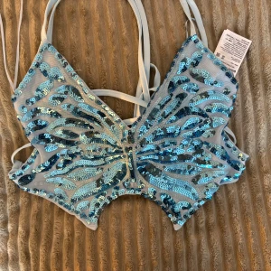 Blå paljettprydd topp - Säljer en glittrig blå topp med paljetter i fjärilsdesign. Toppen har smala axelband och en unik form som ger en iögonfallande look. Perfekt för en festlig kväll!