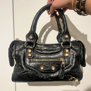 Svart handväska med nitar - Snygg svart handväska i skinn med guldfärgade nitar och dragkedja. Perfekt för att ge din outfit en edgy touch. Väskan har dubbla handtag och flera fack för enkel organisering.