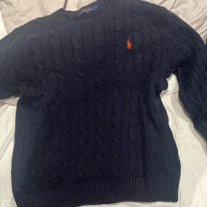 Mörkblå stickad tröja från Ralph Lauren - Säljer en snygg mörkblå stickad tröja från Ralph Lauren med kabelstickat mönster. Tröjan har en klassisk rund halsringning och det ikoniska Ralph Lauren-logotypen i orange på bröstet. Perfekt för kyliga dagar!