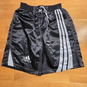 Svarta shorts från Adidas - Säljer ett par svarta Adidas shorts med vita ränder på sidorna och en justerbar snörning i midjan. Perfekta för sport och träning.