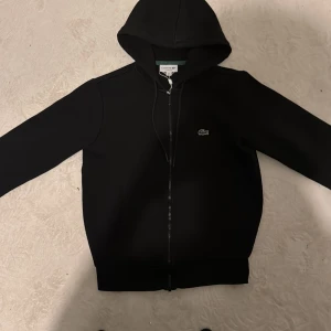 Svart hoodie från Lacoste - Säljer en stilren svart hoodie från Lacoste med dragkedja och klassisk krokodillogga på bröstet. Tröjan har en huva med dragsko och långa ärmar. Perfekt för en avslappnad look.