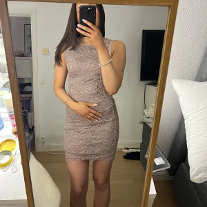 Beige/lika spetsklänning från Bubbleroom - Elegant beige spetsklänning från Bubbleroom med blommigt mönster. Klänningen är ärmlös och har en figurnära passform som framhäver silhuetten. Perfekt för speciella tillfällen.