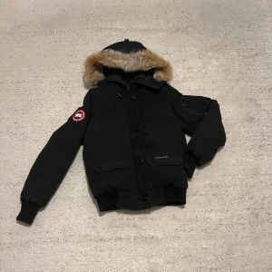 Canada Goose jacka - Säljer en svart dunjacka från Canada Goose med pälsdetaljer på huvan. Jackan har långa ärmar, dragkedja och knappar framtill samt flera praktiska fickor. Perfekt för kalla vinterdagar.