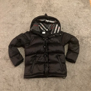 Burberry Puffer jacket (+väst) - Snygg svart dunjacka från Burberry med huva och dragkedja. Jackan har ett klassiskt rutigt mönster på insidan av huvan och är perfekt för kyliga dagar. Den är långärmad och har praktiska fickor med dragkedja. Blir till en väst ochså 