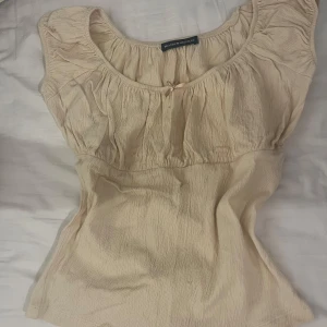 Beige milkmaid topp från Brandy Melville - Säljer en söt beige topp från Brandy Melville med rund halsringning och liten rosett framtill. Toppen har en lätt rynkad design och är ärmlös. Har sytt på rosetten själv. Aldrig använd 
