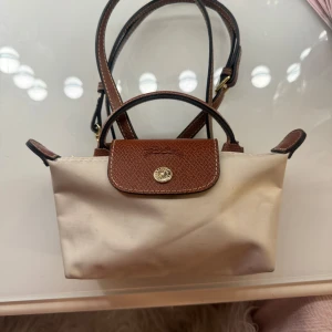 Beige och brun axelväska från Longchamp - Säljer en stilren beige axelväska från Longchamp med bruna skinnremmar och detaljer. Väskan har en praktisk knappstängning och en elegant design som passar perfekt för vardagsbruk. Den är lätt att bära och har en tidlös look. Har ej kvar kvitto 