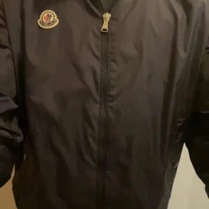  vindjacka från Moncler - Snygg svart vindjacka från Moncler med dragkedja och logotyp på bröstet. Jackan har en huva med Moncler-tryck och röda detaljer. Perfekt för blåsiga dagar.