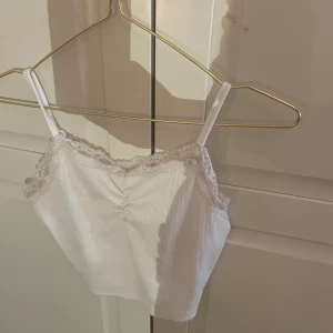 Vit ribbad linnetopp med spetsdetaljer - Söt vit ribbad linnetopp med smala axelband och spetsdetaljer längs kanten. Perfekt för varma sommardagar eller som ett lager under en tröja. Enkel och stilren design som passar till det mesta.