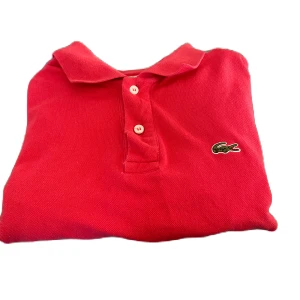 Röd pikétröja från Lacoste - Snygg röd pikétröja från Lacoste med klassisk krage och knappar framtill. Den ikoniska krokodilloggan är broderad på bröstet. Perfekt för en stilren och avslappnad look.