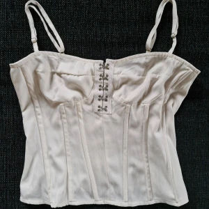 Crèmefärgad korsett från PrettyLittleThing - Snygg crèmefärgad korsett-topp från PrettyLittleThing! Med justerbara axelband och dekorativa hakar framtill. Den har en dragkedja bak för enkel påklädning. Perfekt för en stilren look.