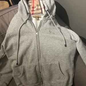 Grå hoodie från Burberry - Säljer en stilren grå hoodie från Burberry med dragkedja och klassiskt rutigt foder i huvan. Tröjan har en broderad logga på bröstet och praktiska fickor framtill. Perfekt för en avslappnad och trendig look.