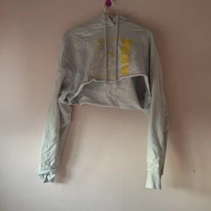 Grå croppad hoodie med tryck - Säljer en grå croppad hoodie med gult tryck framtill. Hoodien har långa ärmar och en justerbar huva. Perfekt för en avslappnad stil.