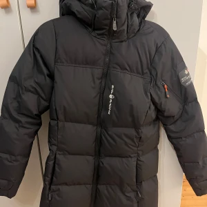 Sail Racing jacka - Snygg och varm svart dunjacka från Sail Racing med huva och dragkedja. Perfekt för kalla dagar med sin långa design och praktiska fickor. Skriv vid fler frågor/bilder🙃