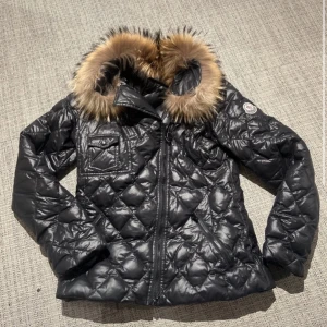 Svart quiltad jacka från Moncler - Snygg svart quiltad jacka från Moncler med pälsdetaljer på huvan. Jackan har en dragkedja framtill och ett Moncler-märke på ärmen. Perfekt för kyliga dagar.
