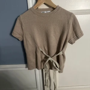 Beige stickad topp från Zara - Säljer en beige stickad topp från Zara med korta ärmar och knytband i midjan. Toppen har en ribbad kant vid halsen och ärmarna. Tröjan är i bra skick och supereftertraktad