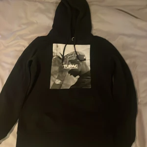 Svart hoodie med Tupac-tryck - Snygg svart hoodie med ett stort tryck av Tupac på framsidan. Perfekt för dig som gillar streetwear och vill ha en bekväm och stilren look. Hoodien har en klassisk passform med långa ärmar och en praktisk huva.