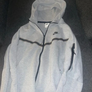 Grå Nike tch fleece - Säljer en grå huvtröja från Nike med dragkedja och svart logotyp på bröstet. Tröjan har en justerbar huva och långa ärmar. Perfekt för en avslappnad stil. Storlek m, bra skick.
