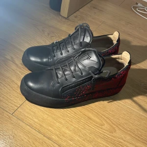 Giuseppe Zanotti Frankie Spray  - Snygga svarta sneakers med röda mönsterdetaljer och dragkedja på sidan. Skorna har snörning och en modern design som passar perfekt för en trendig look. Skolan är i bra skick använd ett par gånger och är bekväma. Inga skador på skorna. Skorna är slutsålda i Zanottis hemsida. Pris går att diskuteras. Boxen finns kvar. 