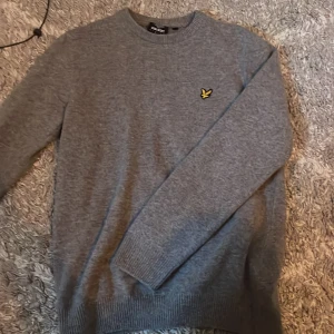 Grå knit från Lyle & Scott - Säljer en stilren grå tröja från Lyle & Scott med deras ikoniska gula logga på bröstet. Tröjan är långärmad och har ribbade muddar vid ärmslut och nederkant. Perfekt för en avslappnad look.