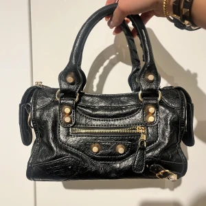 Svart handväska med nitar - Snygg svart handväska i skinn med guldfärgade nitar och dragkedja. Perfekt för att ge din outfit en edgy touch. Väskan har dubbla handtag och en praktisk storlek för vardagsbruk.