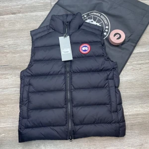 Svart dunväst från Canada Goose - Säljer en stilren svart dunväst från Canada Goose med dragkedja och det ikoniska märket på bröstet. Perfekt för kyliga dagar när du vill hålla värmen utan att kompromissa med stilen. Västen har en hög krage och är quiltad för extra komfort.
