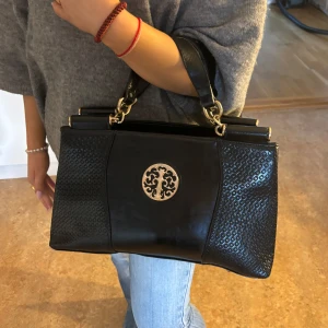 Svart handväska - Snygg svart handväska i skinn från Hasel med ett elegant guldfärgat emblem på framsidan. Väskan har två handtag och en praktisk dragkedja för säker förvaring. Perfekt för att ge en stilren touch till din outfit.