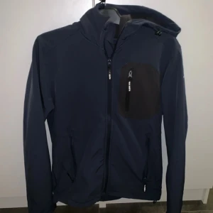 Mörkblå fleecejacka med huva - Snygg mörkblå vindjacka med huva och dragkedja framtill. Jackan har en praktisk bröstficka med dragkedja och justerbara snören vid huvan. Perfekt för blåsiga dagar!