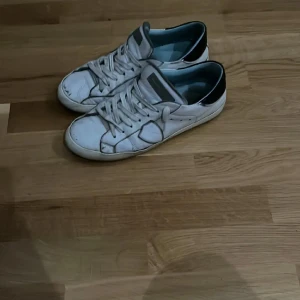 Vita sneakers från Philippe Model - Säljer ett par vita sneakers från Philippe Model med svarta detaljer. Skorna har snörning och en klassisk design. Perfekta för en avslappnad stil.