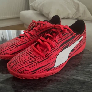 Röda fotbollsskor med svarta ränder - Snygga röda fotbollsskor med svarta ränder och vit logga på sidan. Skorna har snörning och en modern design som passar perfekt för fotbollsplanen.