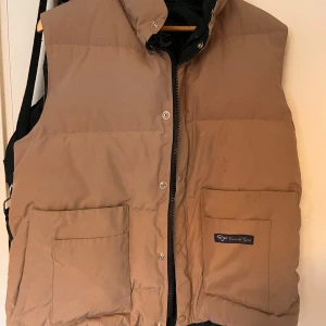 Beige dunväst från Canada Goose - Säljer en beige dunväst från Canada Goose med dragkedja och knappar. Västen har två stora fickor framtill och en hög krage för extra värme. Perfekt för kyliga dagar!