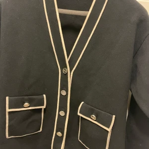 Svart kofta från Sandro - Elegant svart kofta från Sandro med kontrasterande beige kantdetaljer och knappar framtill. Två praktiska fickor med knappstängning ger en stilren look. Perfekt för en chic och sofistikerad stil.