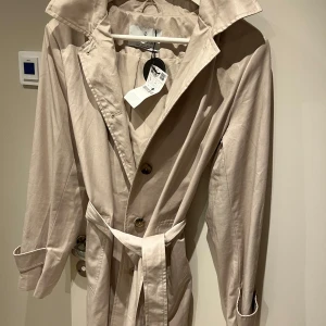 Beige trenchcoat med bälte - Beige trenchcoat med knappar och bälte i midjan. Regnjacka. Liten i storlek.