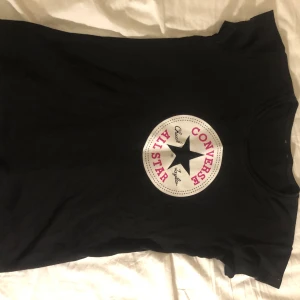 Svart Converse T-shirt - Snygg svart T-shirt med det klassiska Converse All Star-trycket i vitt och rosa på bröstet. Tillverkad av mjuk bomull från Atlant, perfekt för en avslappnad stil.