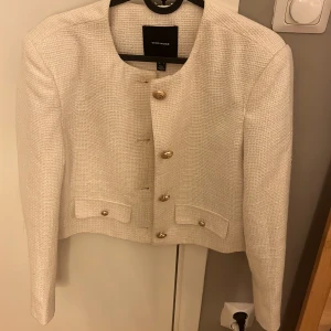 Vit kavaj/kofta från Vero Moda - Säljer en stilren vit kavaj/koftan från Vero Moda med guldknappar och två fickor framtill. Kavajen/koftan har en croppad passform och långa ärmar, perfekt för en chic look.