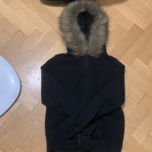 Svart jacka med pälskrage - Säljer en svart jacka med huva och pälskrage. Jackan har dragkedja framtill och långa ärmar. Perfekt för kyligare dagar.